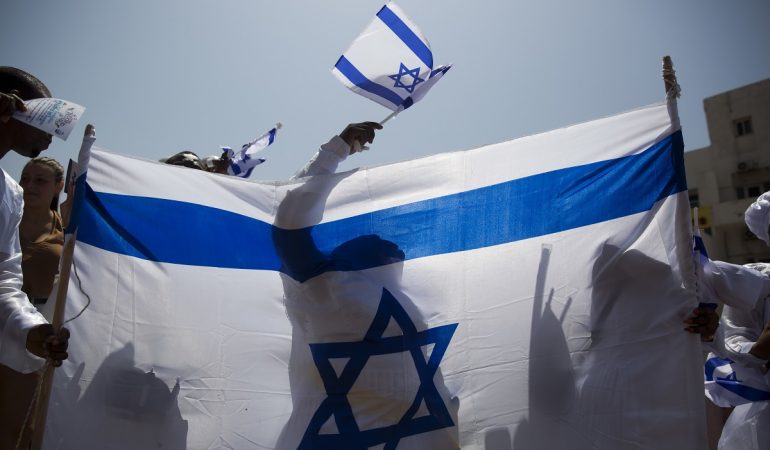 19.04.2018, Israel, Tel Aviv: Ein Feiernder schwenkt eine israelische Flagge hinter einer großen Fahne am 70. Unabhängigkeitstag des jüdischen Staates.